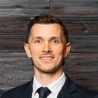 Chiropractor John Pettygrove In Suit Smiling In Front Of Wooden Wall